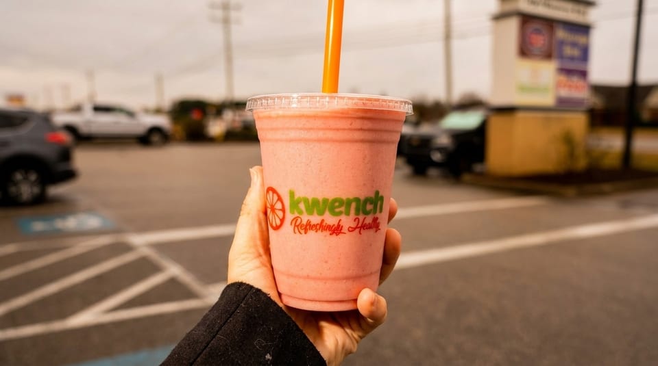 Hand holding a Kwench smoothie with an orange straw, outdoors in a parking lot.