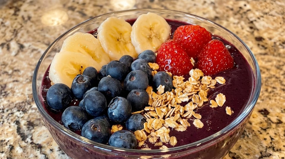 Acai bowl topped with sliced bananas, blueberries, strawberries, and granola in a clear bowl.