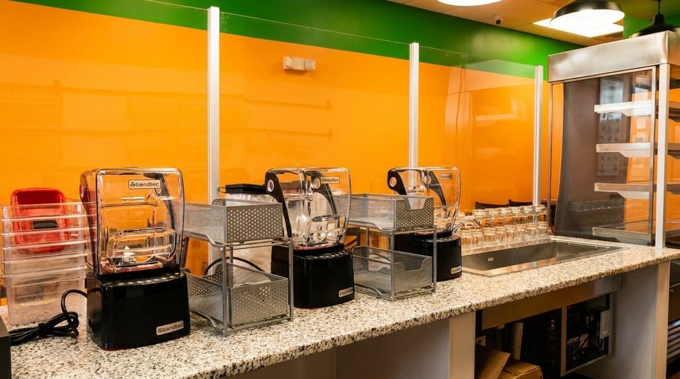 Three Blendtec blenders and metal baskets on a granite counter with an orange and green wall.