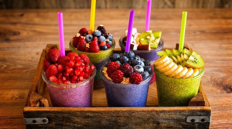Five colorful chia puddings with fresh fruit toppings and straws in a wooden tray.